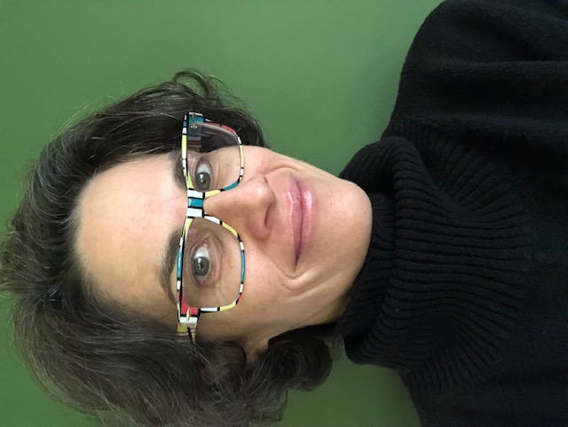 professional headshot of Kelly in Mondrian inspired eyeglasses, a black turtleneck sweater, with a forest green background